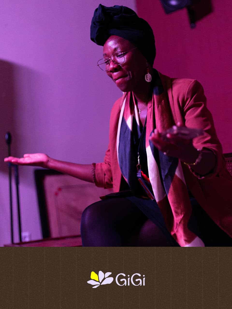 A storyteller in a headwrap and glasses, smiling, one hand open as she speaks from the chair in pink light.